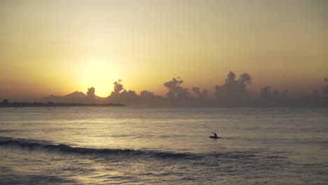 A-man-on-a-small-traditional-boat-sailing-towards-the-sea-at-sunset-or-sunrise-with-dramatic-clouds-in-Bali,-Indonesia,-Slow-motion