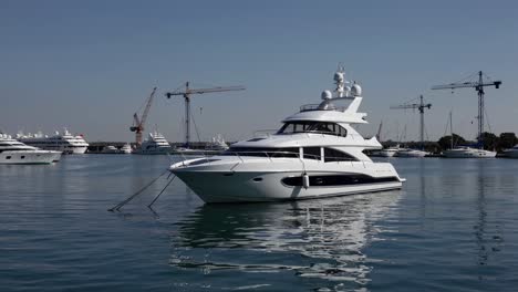 luxurious white yacht floating near marina, reflecting cloudless sky with industrial cranes silhouetted in background, suggesting urban waterfront landscape