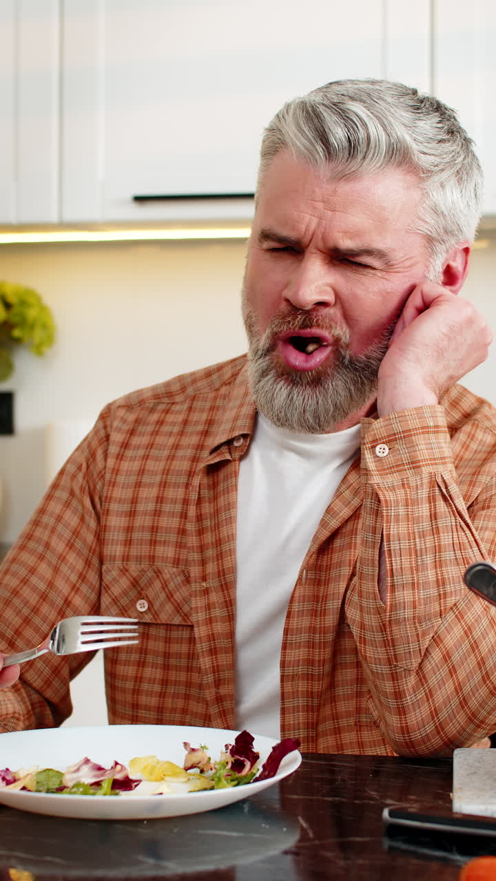 Middleaged man grimacing while eating healthy salad forcing diet willpower in modern home kitchen