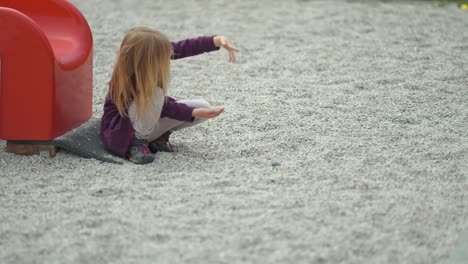 Ein-Junges-Blondes-Mädchen-Sitzt-Allein-Neben-Der-Rutsche-Auf-Dem-Spielplatz-Im-Freien-Und-Spielt-Mit-Steinen