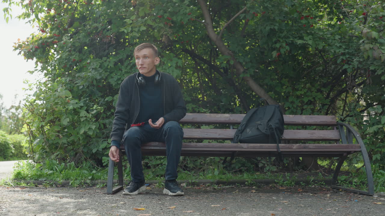 Giovane seduto tranquillamente con gadget, giovane in stile casual che si gode un tranquillo ambiente urbano all'aperto, giovane con cuffie e zaino che trova la calma in un ambiente urbano naturale