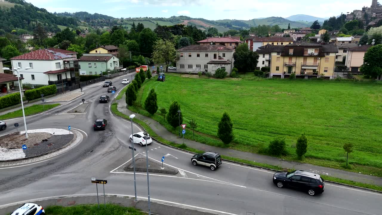 Drone view View of a Blue Tractor Mowing Green Grass Along a Roadside with a White Car and Houses in the Background on a Cloudy Day