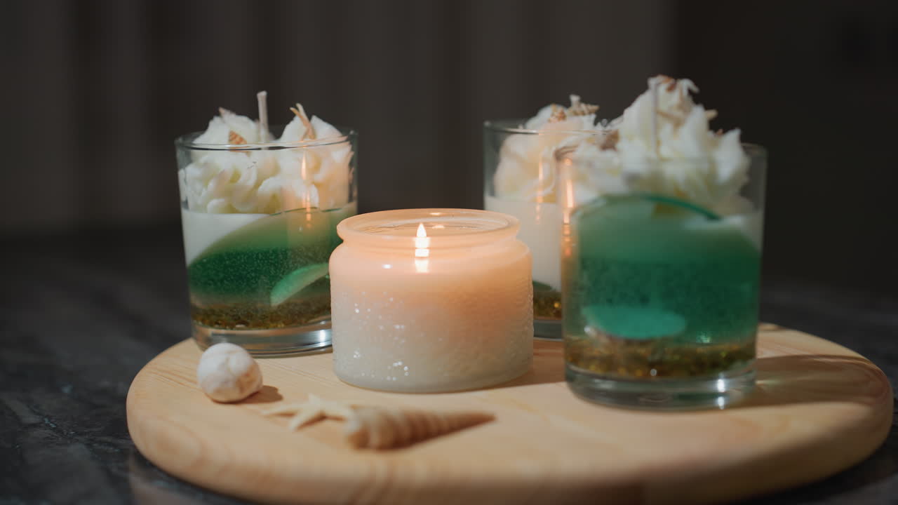 Round view of beautifully arranged candle containers on wooden tray, one candle lighted casting soft glow and gentle reflection on nearby unlit decorative candles surrounded by elegant cream swirls