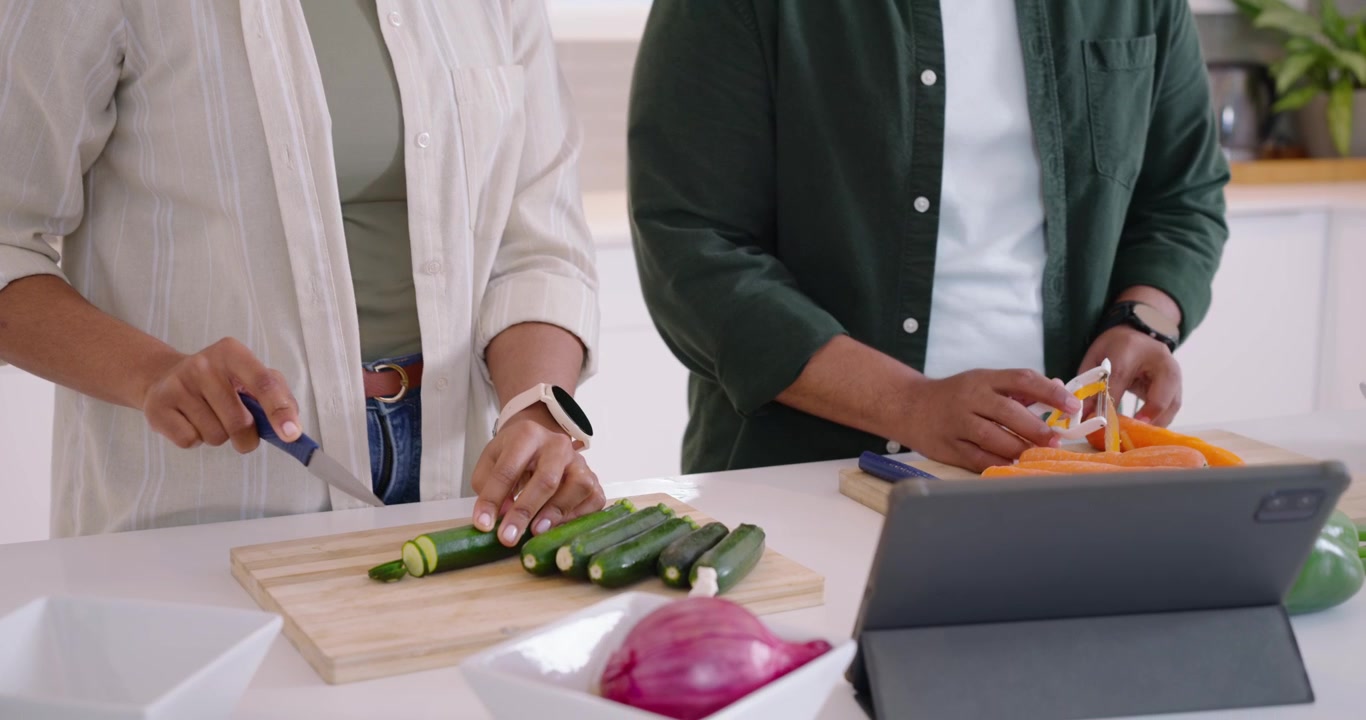 Coppia afroamericana che usa una ricetta su tablet al bancone, la donna affetta le zucchine e l'uomo pela le carote