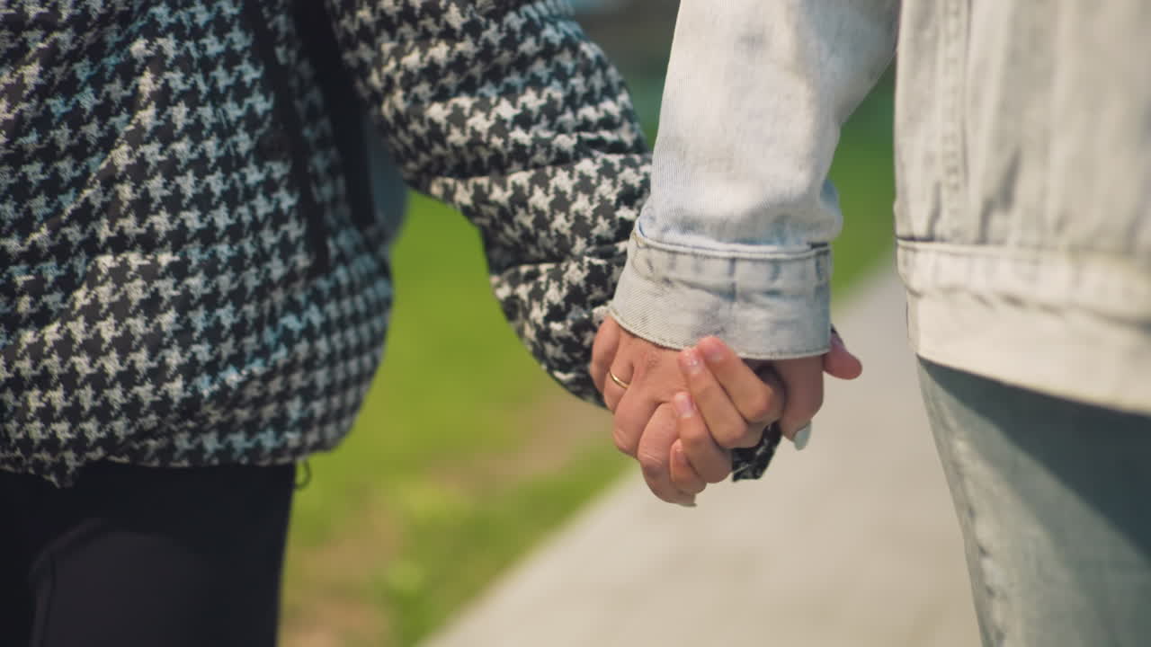 Close-up of hands gently holding on city sidewalk, one wearing checkered coat and the other denim jacket, symbolizing care, support, and trust between two people in warm, emotional moment during urban walk