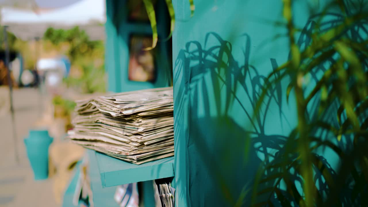 Bancarella di libri all'aperto con un muro blu calypso e una grande pila di giornali nell'angolo. Inquadratura generale statica in slow motion durante una mattina soleggiata nel Sindh, Pakistan, con profondità di campo ridotta.