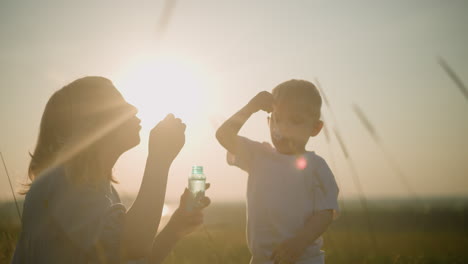 a woman in a blue dress bends in a grassy field at sunset, holding a bubble bottle as she and her young son, dressed in a white shirt, blow bubbles from wands. the sun's rays create a glowing effect