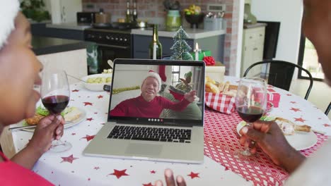 African-american-couple-with-wine-using-laptop-for-christmas-video-call-with-man-on-screen