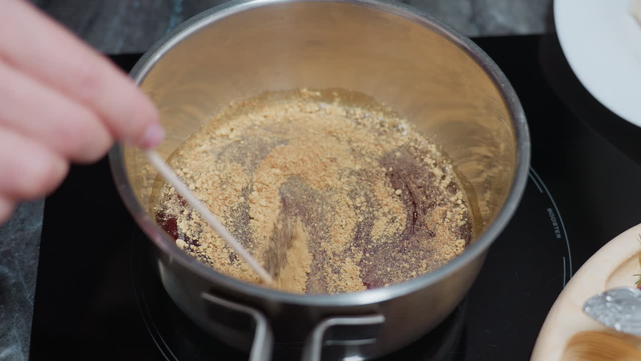 Primo piano di una mano che usa un bastoncino per mescolare la zuppa in una pentola d'acciaio inossidabile su un piano cottura elettrico, con ingredienti in polvere che si mescolano nel liquido, un vassoio di legno e utensili da cucina posizionati nelle vicinanze