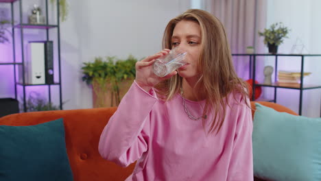 Thirsty-one-young-woman-sitting-at-home-holding-glass-of-natural-aqua-make-sips-drinking-still-water