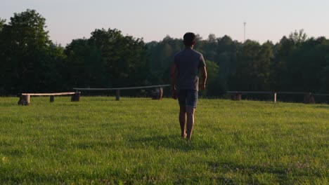 Rear-view-of-young-man-walking-away-from-camera-on-park-meadow,-golden-hour