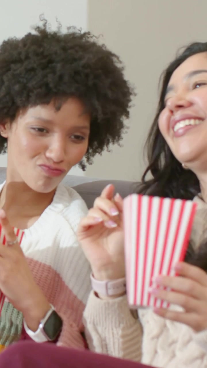 Video verticale: Due donne sul divano che reagiscono alla TV, condividendo popcorn, una con un top a righe e l'altra con una maglia color crema.