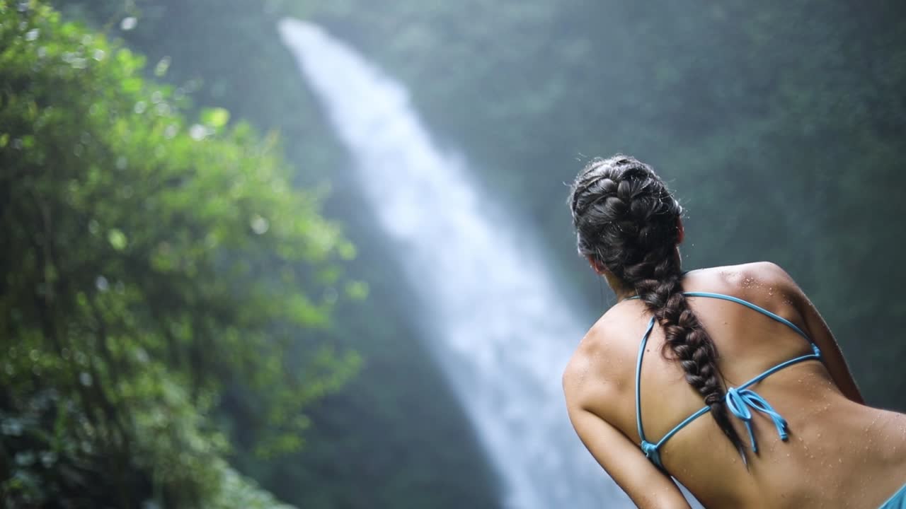 toma panorámica en cámara lenta de una chica en bikini azul sentada frente a una cascada de nungnung en bali, indonesia