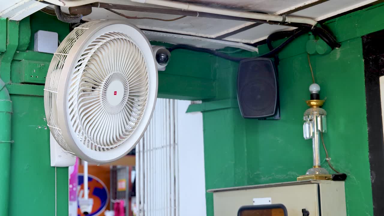 White wall-mounted fan oscillates in sheltered outdoor urban area with green walls and daylight