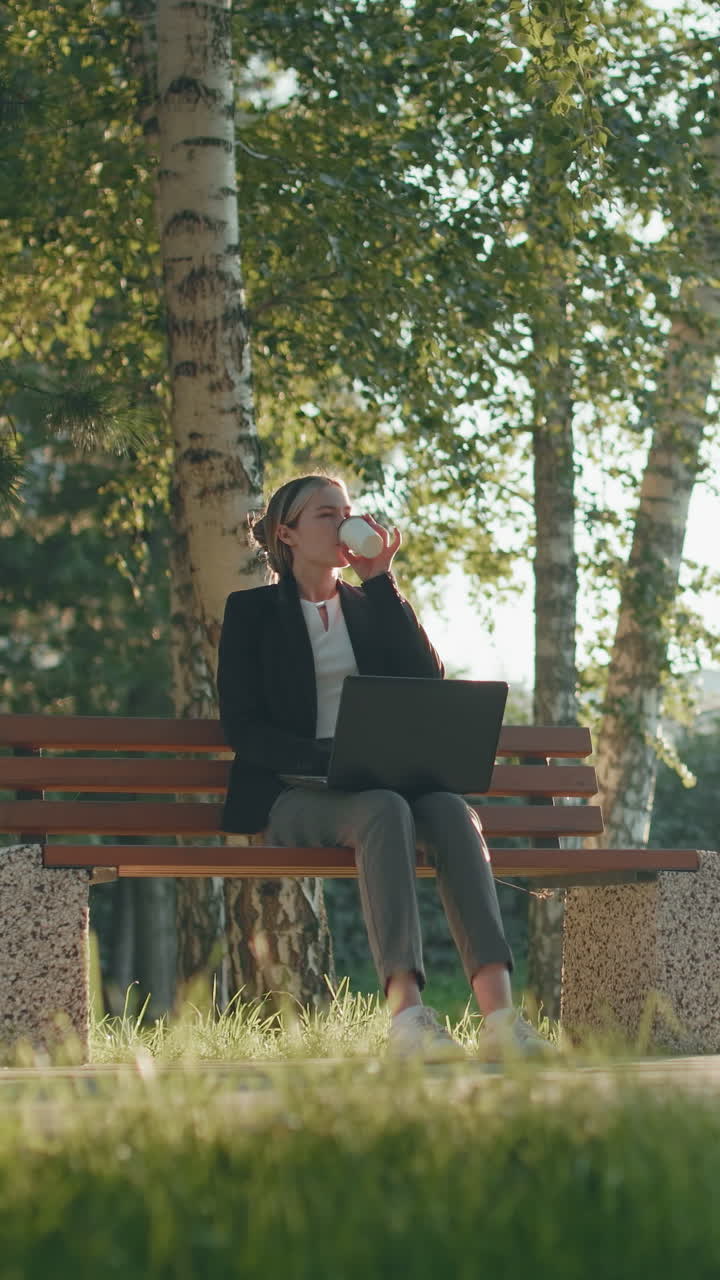Freelancer seduto all'aperto su una panchina di legno mentre lavora sul sistema e sorseggia un caffè, con alberi verdi, luce solare mattutina e un autobus in lontananza in un parco urbano
