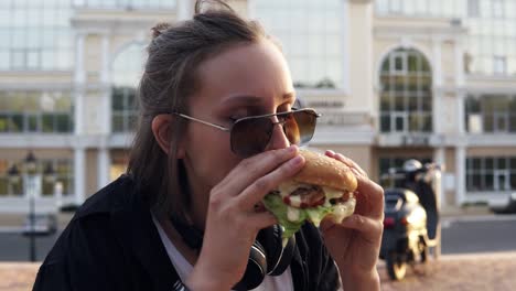 young female eats a big, juicy hamburger with two hands. young girl with short hair and dark sunglasses. hungry. close up