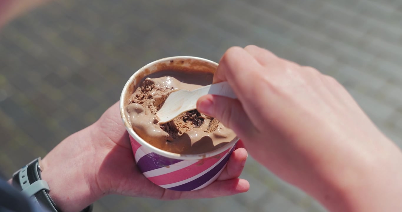 Primo piano delle mani di un bambino che mangia gelato al cioccolato da una coppetta all'aperto in una giornata di sole.