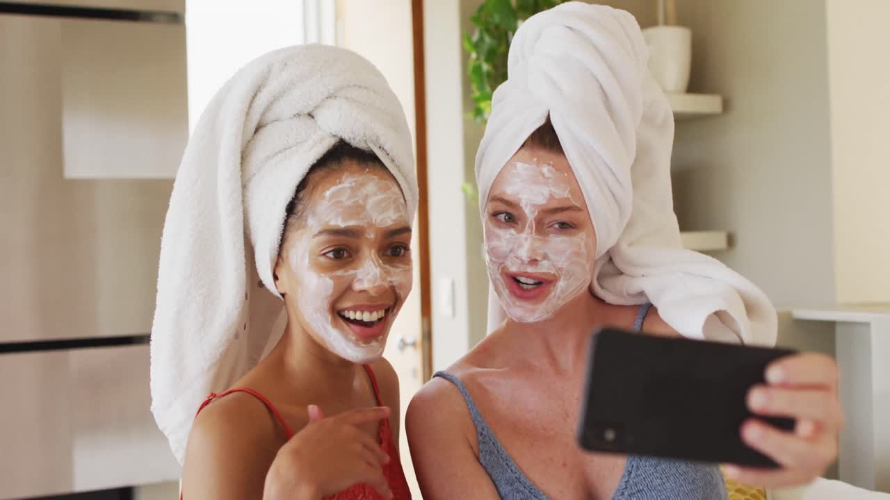 Diverse amiche felici con asciugamani in testa e maschere per il viso che si scattano un selfie a casa