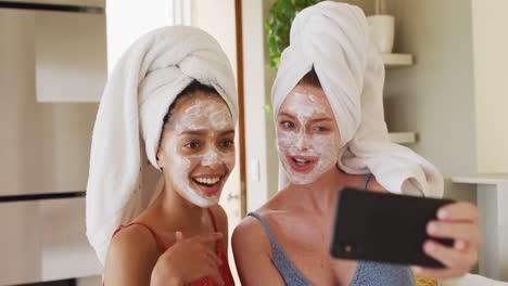 Diverse-happy-female-friends-wearing-towels-on-heads-and-cleansing-masks-taking-selfie-at-home