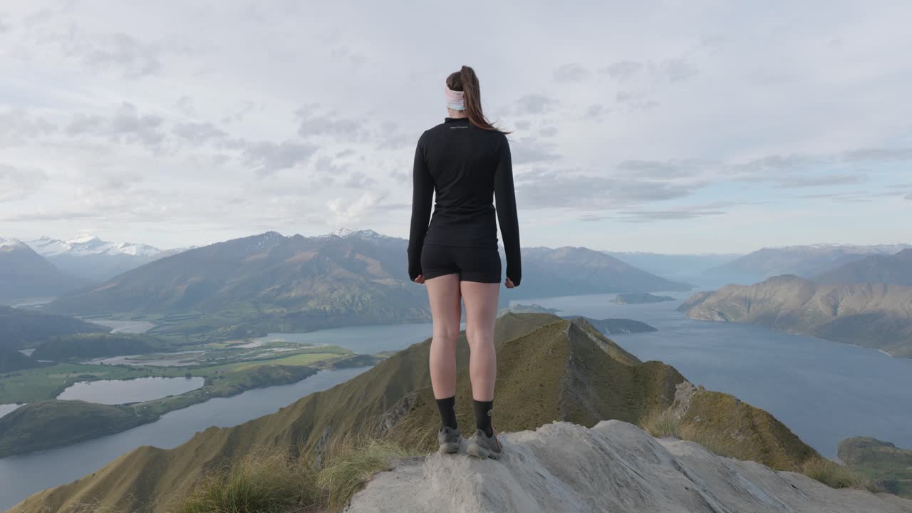 joven mujer caucásica con cola de caballo y en pantalones cortos de pie en el punto de vista en el pico de roy, mirando el lago wanaka y las montañas en wanaka, nueva zelanda