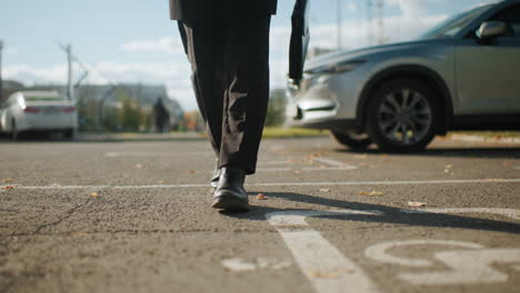 leg view of man in black walking confidently past parked cars on urban pavement under sunlight, carrying a briefcase, surrounded by autumn leaves, with blur view of people walking in the background