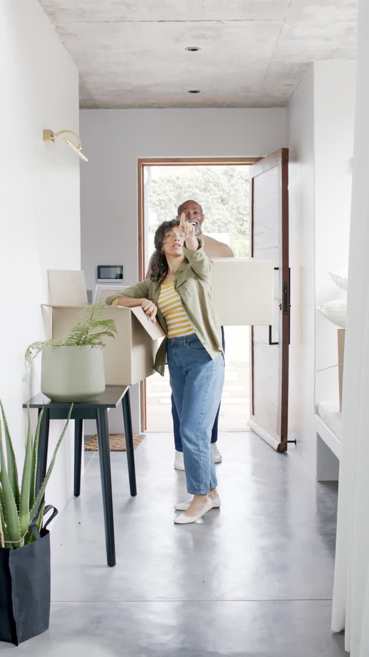 Vertical video of happy diverse mature couple moving into home carrying boxes, slow motion