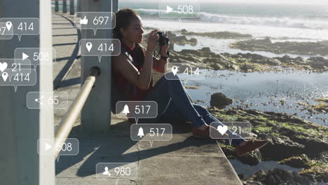 social media notifications animation over woman sitting by ocean taking photos