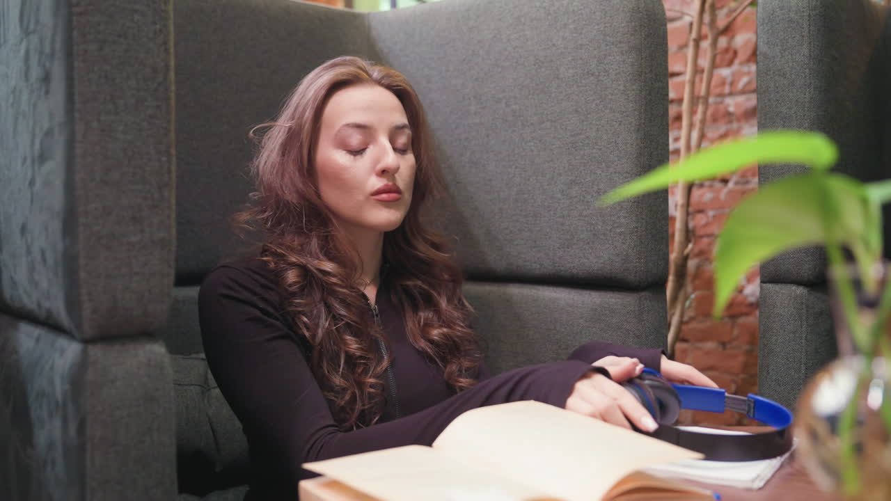 Light skin lady with blond hair taking off black headphones while sitting in cozy booth, looking thoughtful with open book and notepad on table, surrounded by warm ambiance and indoor plants