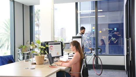 working in modern office, woman using laptop while man with bicycle in background