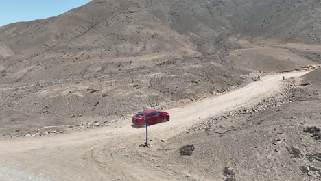 Toma-Panorámica-Aérea-De-Un-Vehículo-Rojo-En-Movimiento-Durante-Un-Viaje-Corto-En-El-País-Desértico-De-Perú-En-Una-Carretera-Polvorienta-Con-Colinas,-Piedras-Y-Montañas-En-Un-Día-Soleado-Sin-Nubes