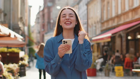mujer joven usar teléfono inteligente móvil celebrando ganar buen mensaje de noticias al aire libre en la calle de la ciudad urbana