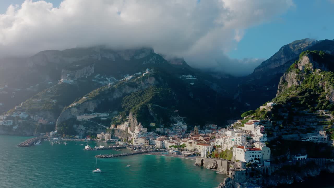 Esclusiva vista aerea con drone di una città costiera della Costiera Amalfitana con edifici storici bianchi, molo per barche, pontile del porto, scogliere imponenti e acque turchesi del Mar Mediterraneo al sole della sera