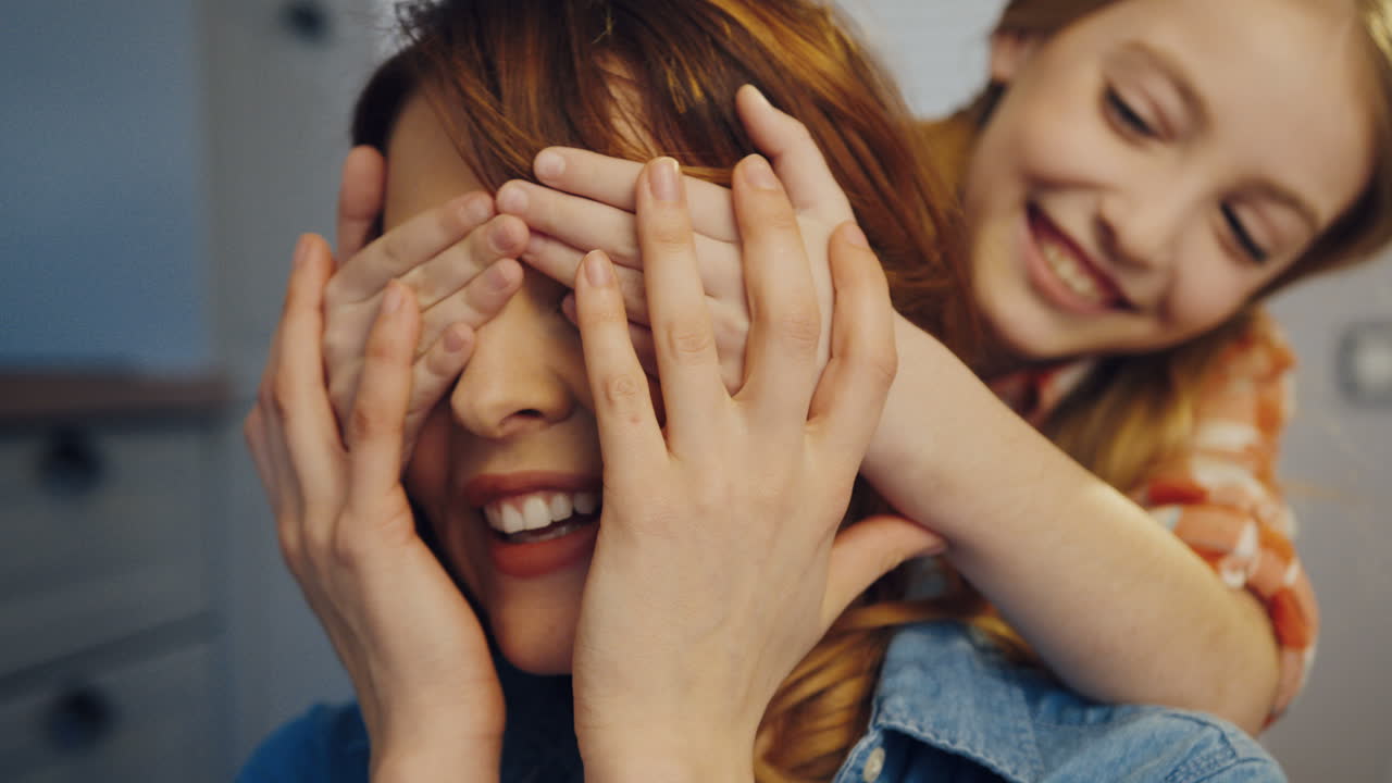 Close up of the beautiful woman, her daughter closing mom's eyes from behind and than they hugging. Portrait shot. Indoors