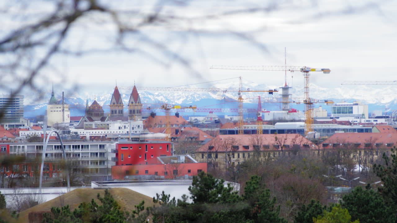 A scenic view of Munich’s skyline with construction cranes and historic buildings, set against the snow-capped Alps. Captured on a cloudy winter day