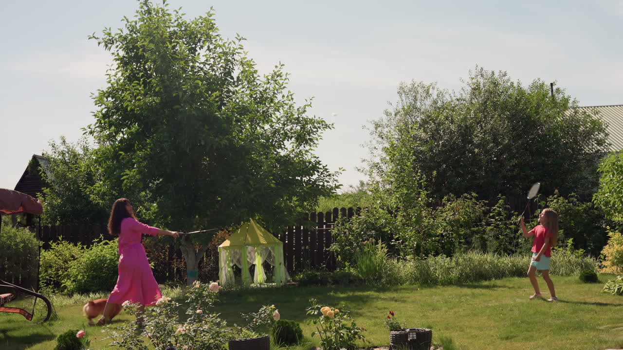 White Mother Guides Daughter Through Garden Beside Tall Tree, Teaching About Plants And Flowers, Green Foliage And BlossomFilled Beds Create Gentle Educational Summer Scene, Curious Child Follows