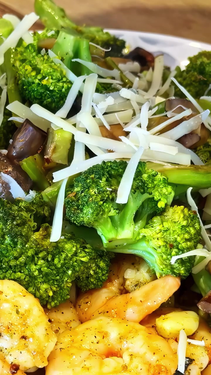 Gamberi cotti e conditi serviti con broccoli e funghi saltati in padella, guarniti con formaggio grattugiato su un piatto bianco. Video verticale, ideale per i social media.