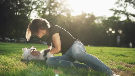 front footage of a woman in jeans and black t shirt lying on the green grass in the park and playing with her pet