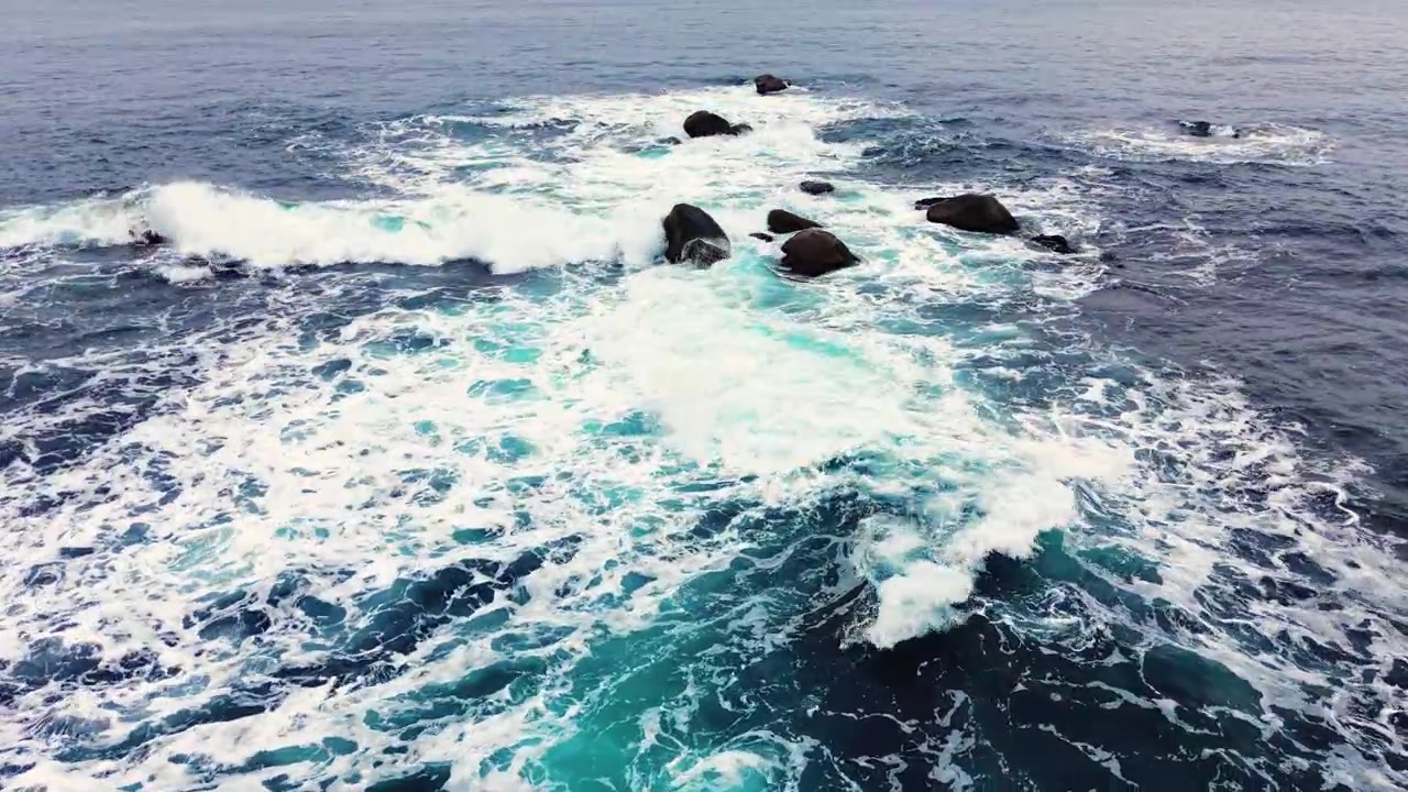 Vista aerea di onde che si infrangono su rocce sparse. Mare blu scuro, schiuma bianca e numerosi massi lungo la costa