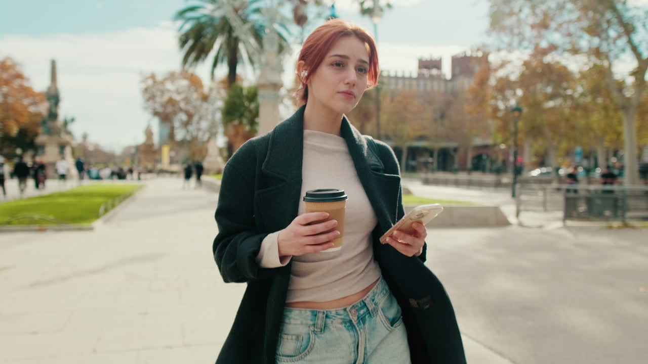 Giovane donna che cammina in un parco cittadino con caffè e smartphone