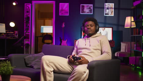 gamer at home sitting down on couch enjoying games on laptop during leisure time. black man sitting on couch in living room, playing video games on notebook, relaxing after work. camera b.