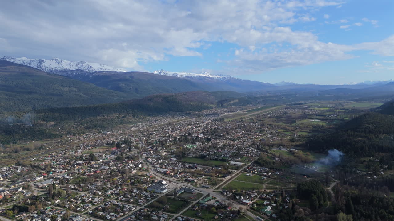 imágenes aéreas de alta resolución de el bolson ubicado en un valle pintoresco, rodeado de majestuosas montañas, bosques y campos verdes bajo un cielo parcialmente nublado.