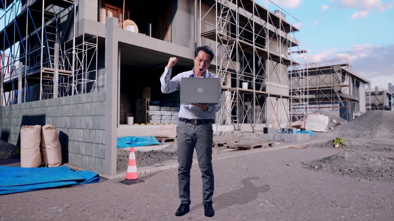 Full Body Of An Asian Male Professional Worker Standing With His Laptop At Construction Site in Progress to New House, He Raises His Fist Up With Screaming Goal