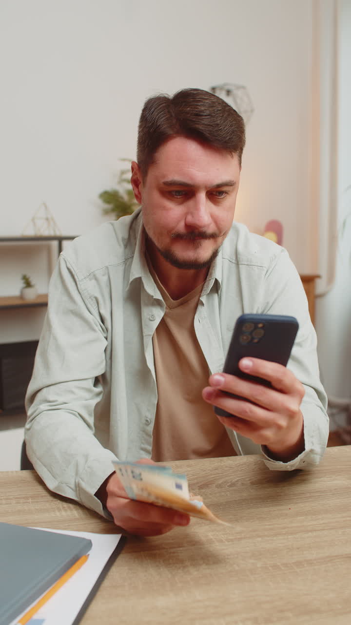 Giovane caucasico sorridente che conta il denaro contanti usando lo smartphone a casa tavolo dell'ufficio in soggiorno