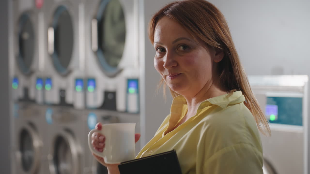 Portrait of female entrepreneur holding notebook while sipping coffee inside modern laundromat, industrial washer and dryer background, stainless steel polished, digital panels glowing
