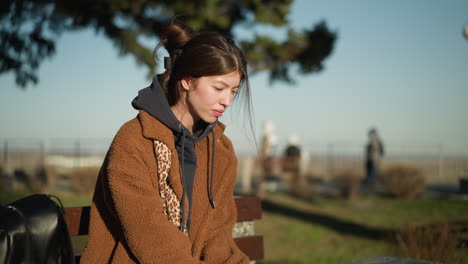 a girl sits alone on a bench in an urban park, looking unfortunate and lost in thought as she gently shakes her head. she is wearing a brown coat, blue jeans, and white shoes