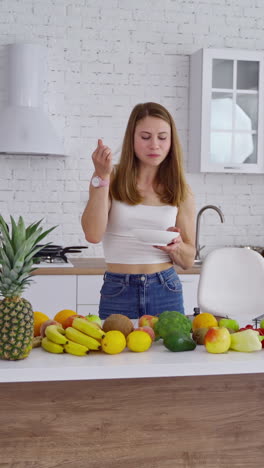 happy young woman in the kitchen. beautiful girl eating something while standing at the table with many organic fruit and vegetables at home. vertical video