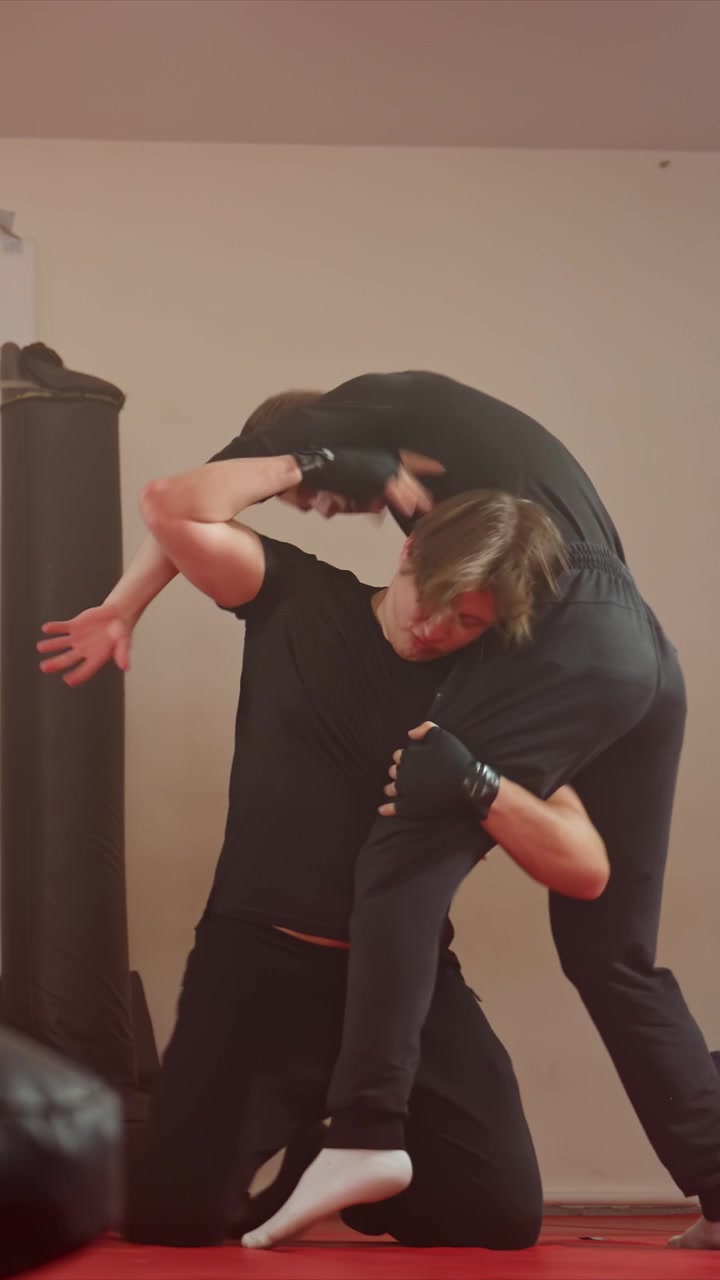 Young man in black clothing adjusts waistband. Second young man joins for grappling demonstration with padded equipment. Plain beige wall background minimal objects