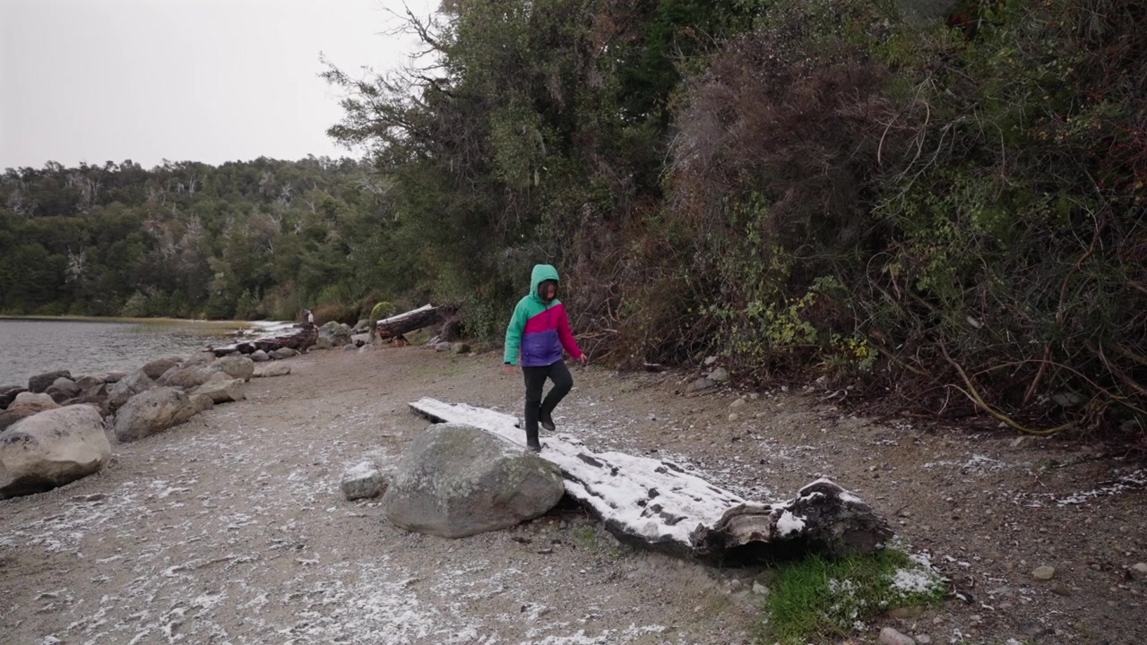 A young child in a colorful jacket balances while walking on a snowcovered log by a lake in Bariloche. Authentic winter exploration and wellness in nature.