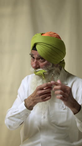 vertical video close up shot of senior sikh man with beard tying fabric for turban and using salai needle against plain background 1