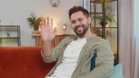 happy young man vlogger smiling friendly at camera and waving hand while sitting on sofa at home